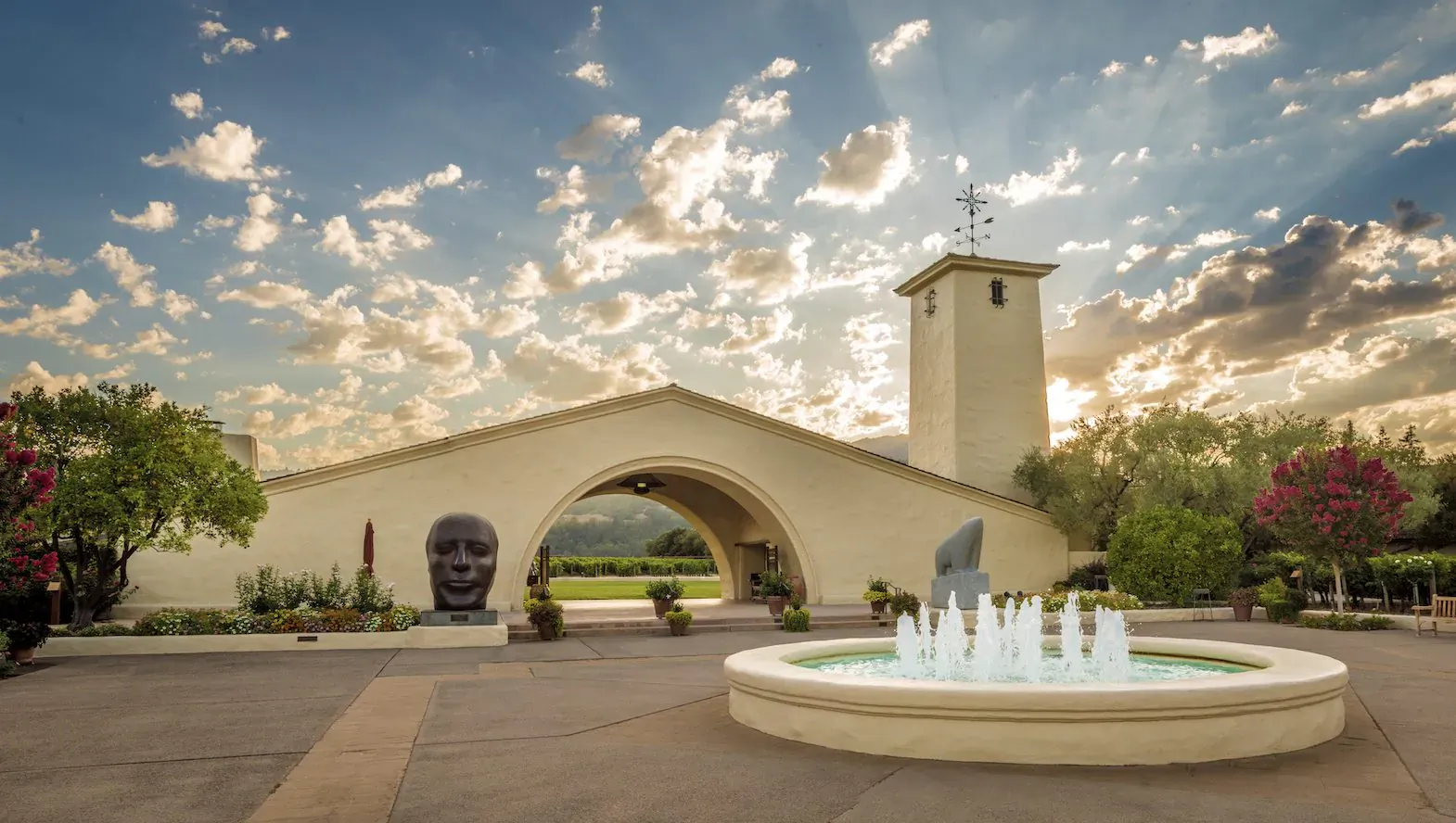 Beaux-Arts winery entrance with arched gateway, bell tower, stone heads, fountain, and vineyard under sunset sky