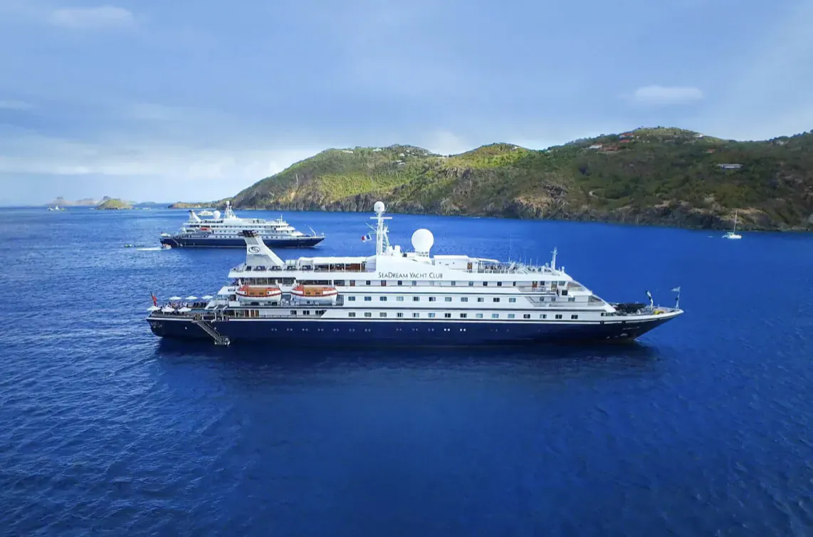 White SeaDream yacht with satellite dish and shore power setup anchored in blue sea near green hills, other yachts visible.