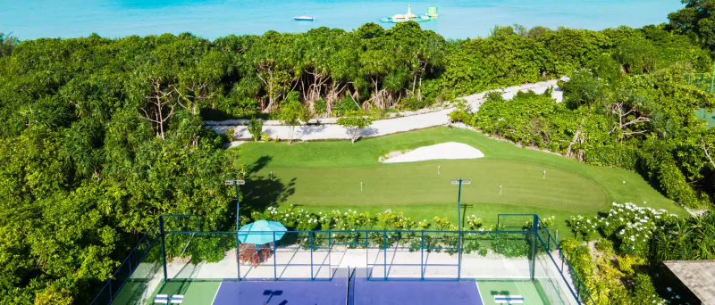 Aerial view of purple tennis court, golf green, lush greenery, and turquoise ocean at Hideaway Beach Resort, Maldives.