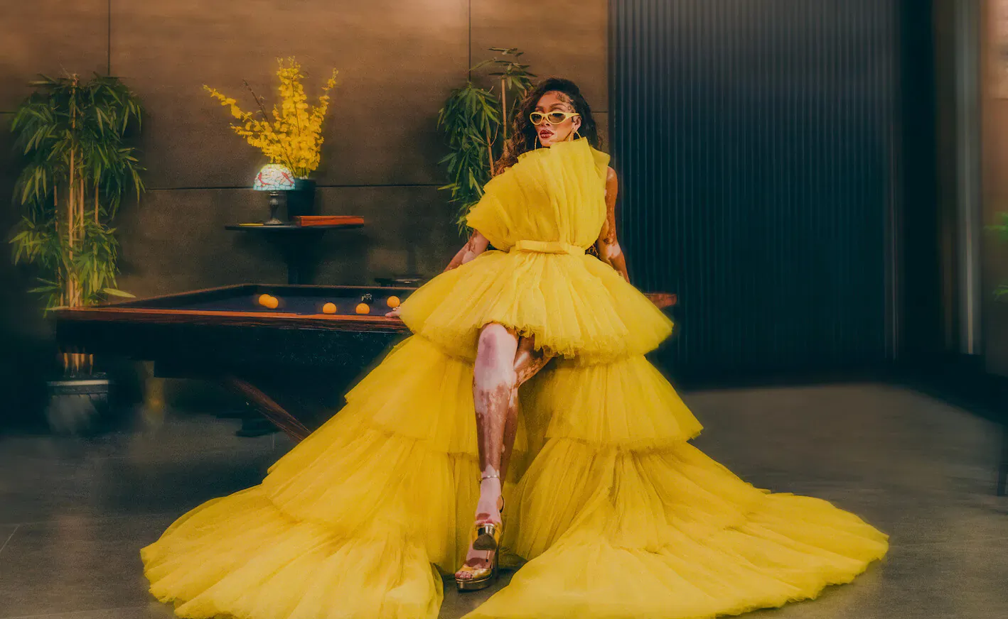 Woman in voluminous yellow tulle gown with high slit, sitting elegantly by table with yellow flowers in modern room