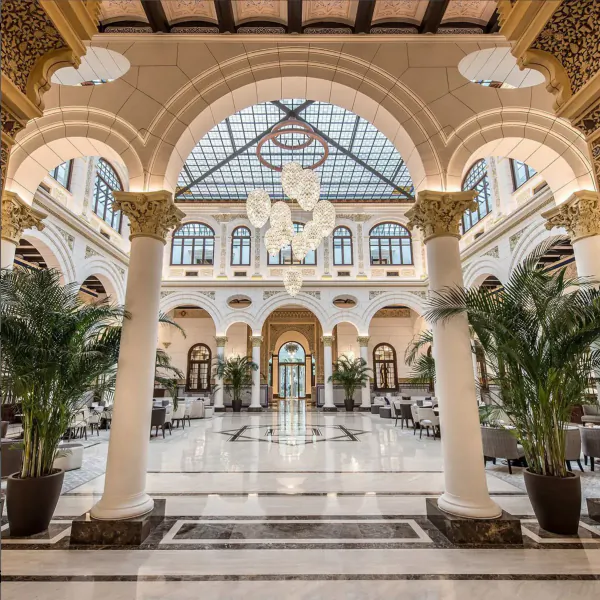 Opulent hotel lobby with arched ceilings, glass dome, chandeliers, marble floors, columns, palms, and seating.