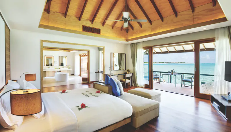 Luxurious Maldives beachfront bedroom with wooden ceiling fan, king bed with rose petals, en-suite bathroom, and ocean view balcony.