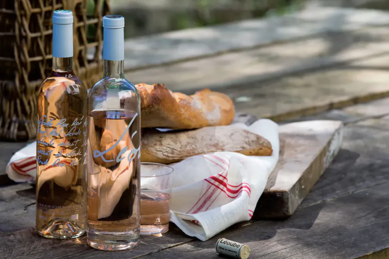 Two Château Léoube rosé wine bottles, baguette bread on cutting board, red napkin in wicker picnic basket on wooden table outdoors.