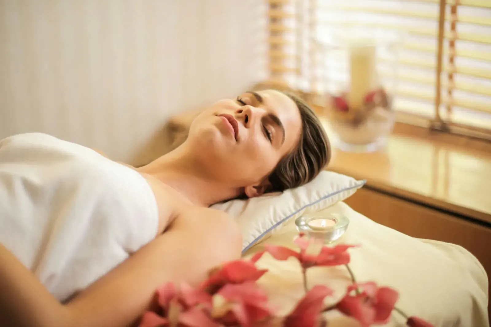 Serene woman lying relaxed on massage table, wrapped in white towel, with pink orchids and candle nearby