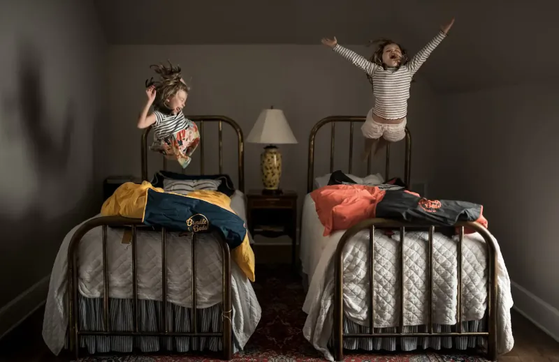 Two joyful girls jumping on twin beds in a cozy bedroom with colorful bedding and lamp.