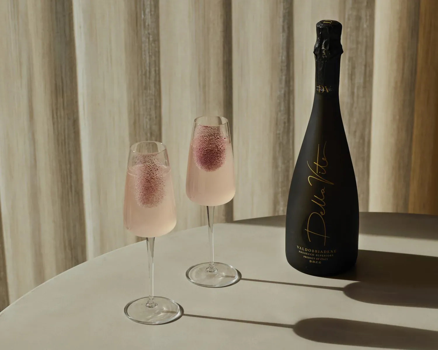 Two pink Paloma Bellini cocktails in flute glasses beside a Della Vite sparkling wine bottle on a table.