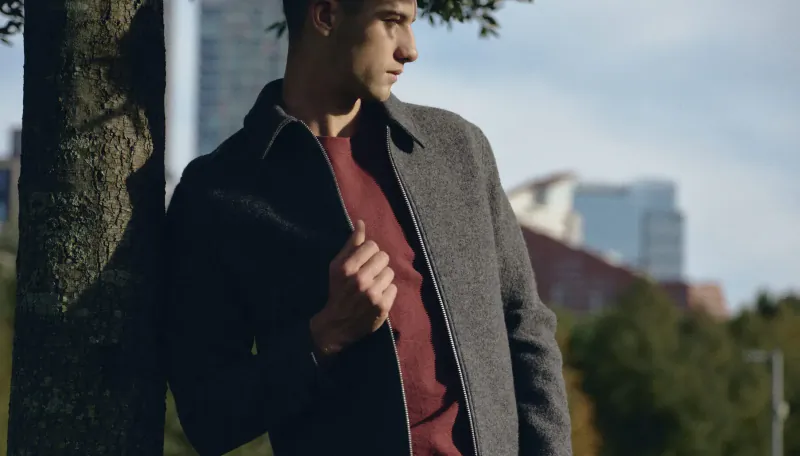 Man in grey coat and red shirt leans against tree in Canary Wharf, urban skyline behind.
