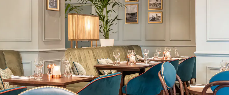 Elegant dining area at Browns Cardiff with teal velvet chairs, wooden tables set with wine glasses and candles, framed art and plants on walls.