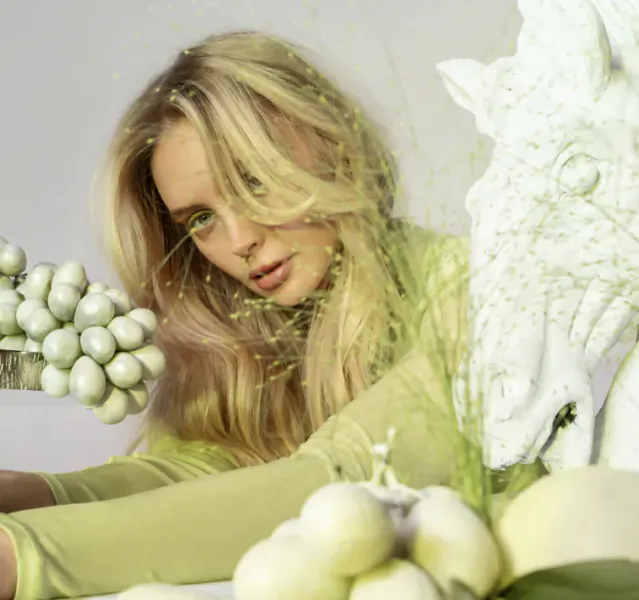 Blonde woman in green top poses with white horse sculpture and white grapes, surrounded by grass