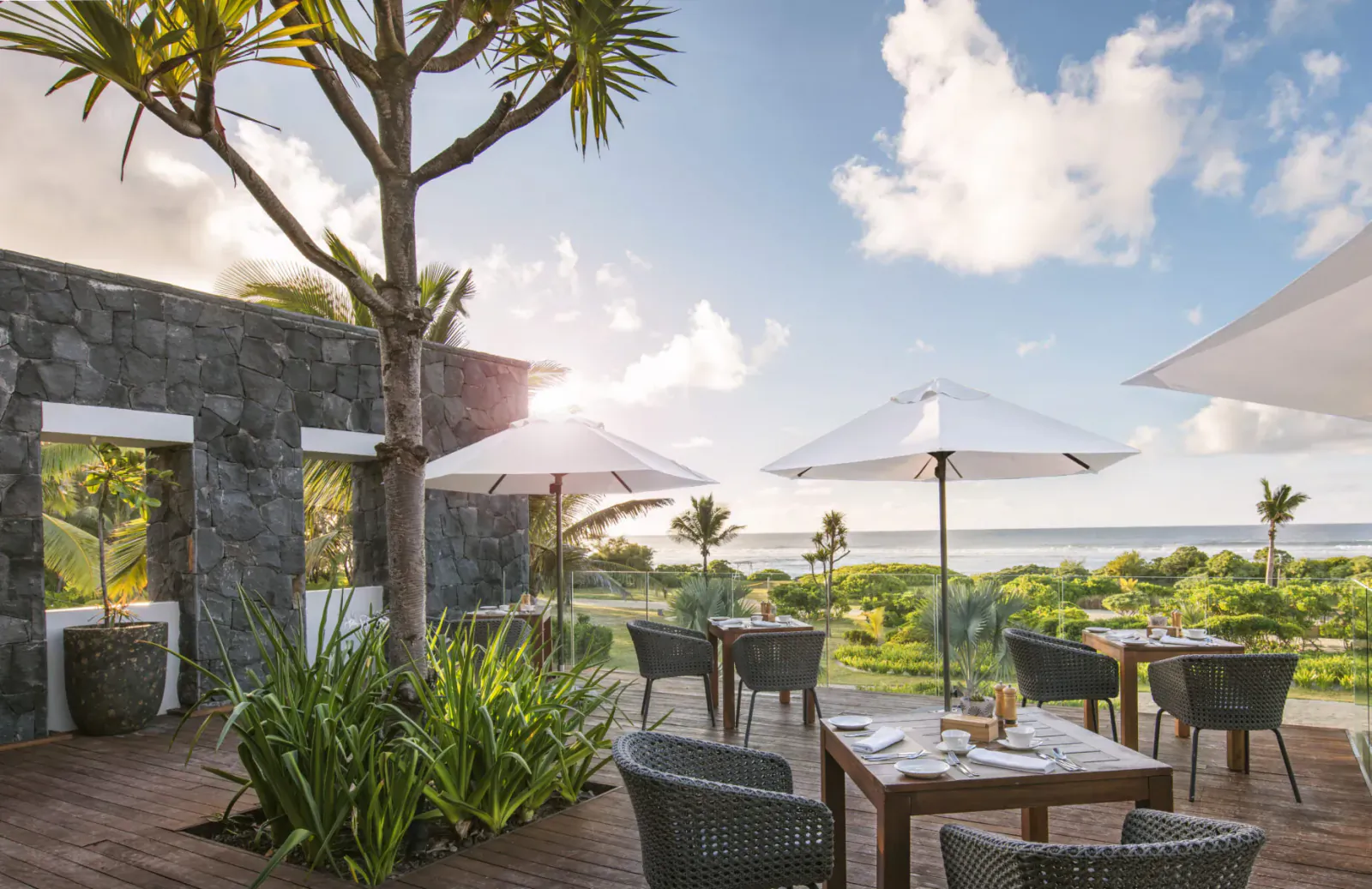 Outdoor terrace at Anantara Iko Mauritius resort with wicker tables, white umbrellas, palm trees, and ocean view.