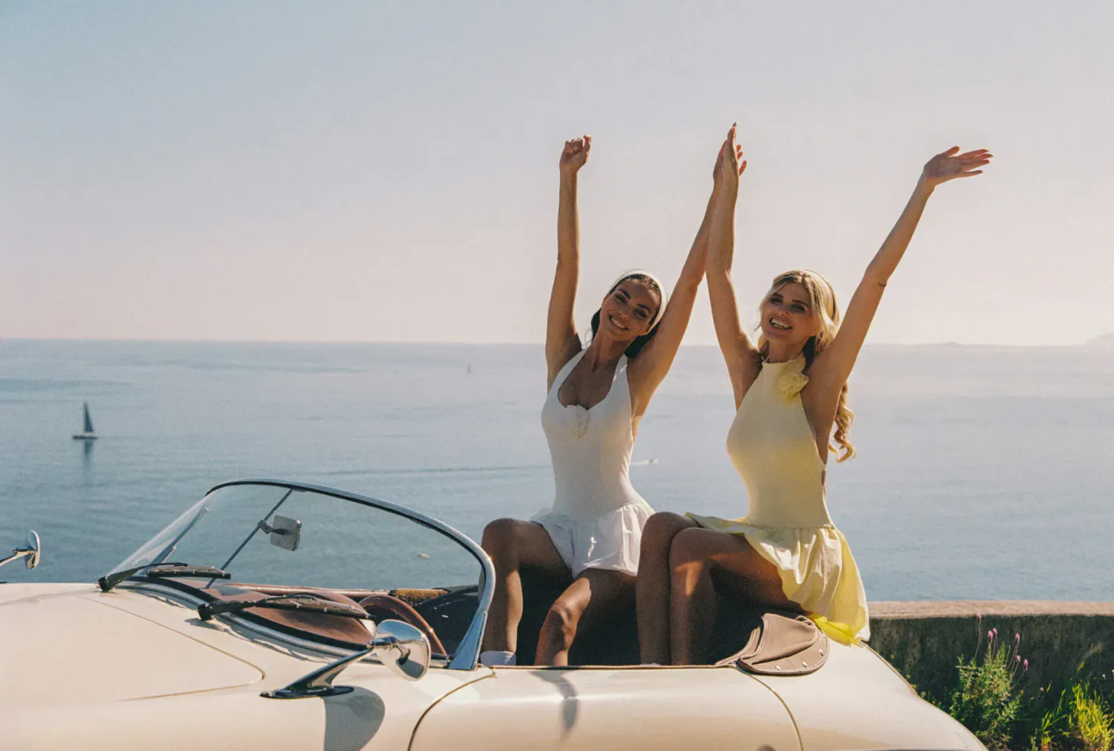 Two women in white and yellow dresses raise arms joyfully, sitting on white convertible overlooking sea and sailboat.
