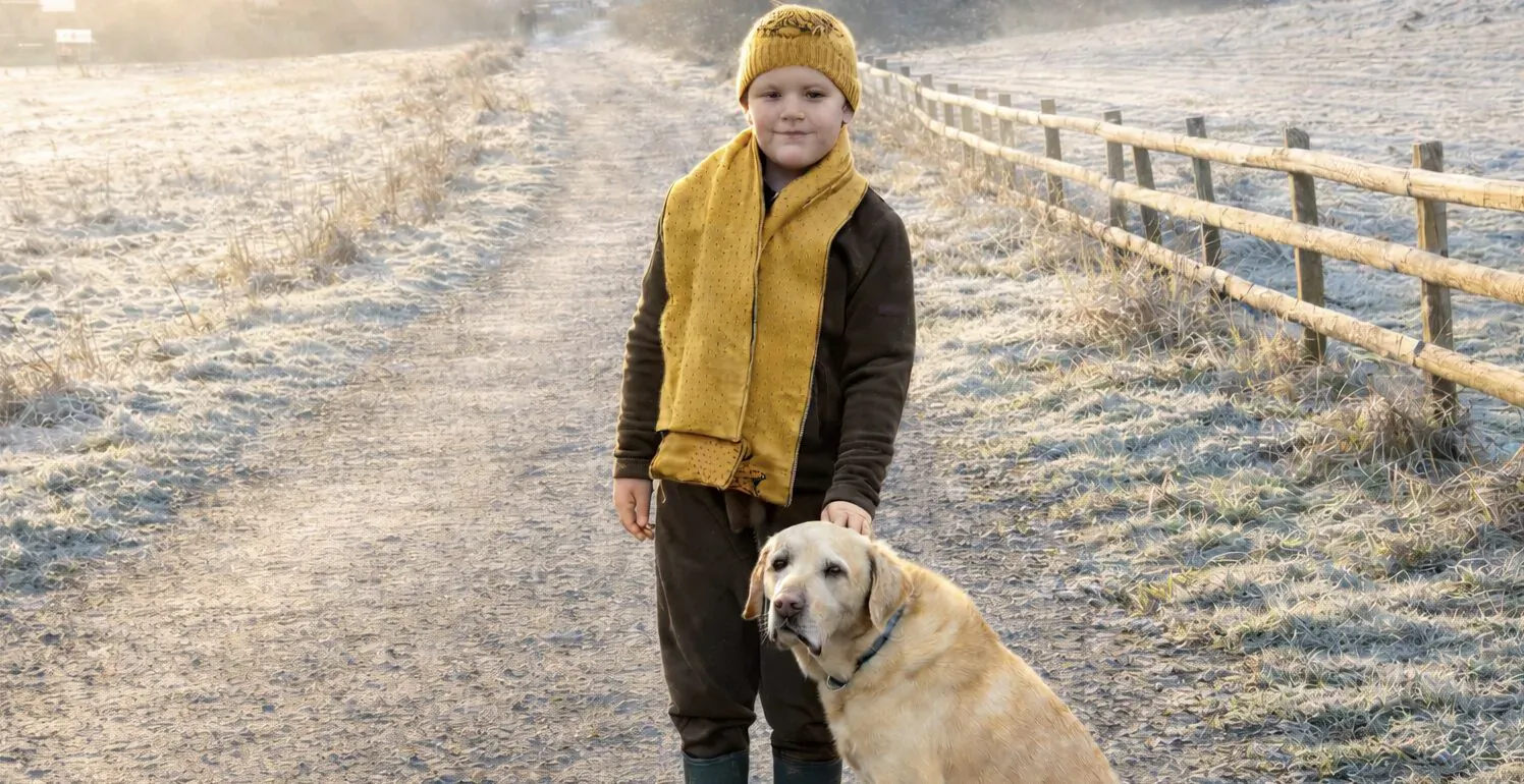 Young boy in yellow knit hat and scarf holds leash of Labrador retriever on frosty rural path.