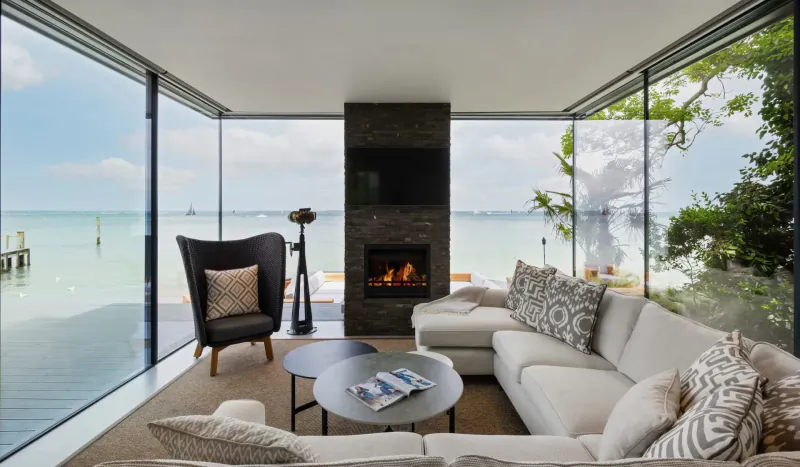 Modern living room with white L-shaped sofa, black armchair, coffee tables, lit fireplace, and panoramic lake views through glass walls.