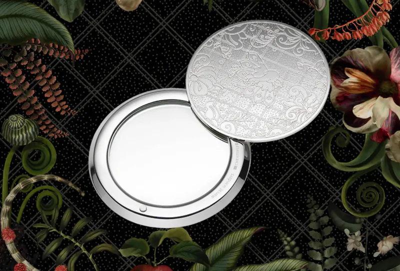 Open engraved silver Jardin d’Éden compact on black checkered background with lush floral motifs