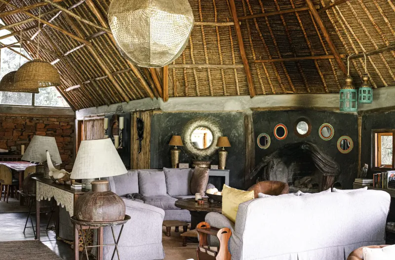 Cozy living room in Kenyan thatched lodge with gray sofas, gold pendant light, mirrors, lamps, and fireplace.