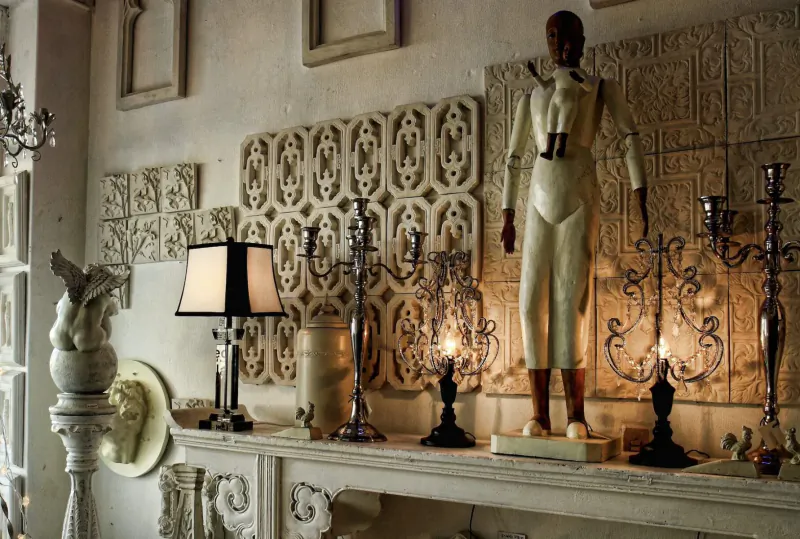Tall white mannequin stands on ornate mantelpiece amid candelabras, lamps, and carved wall panels in gallery.