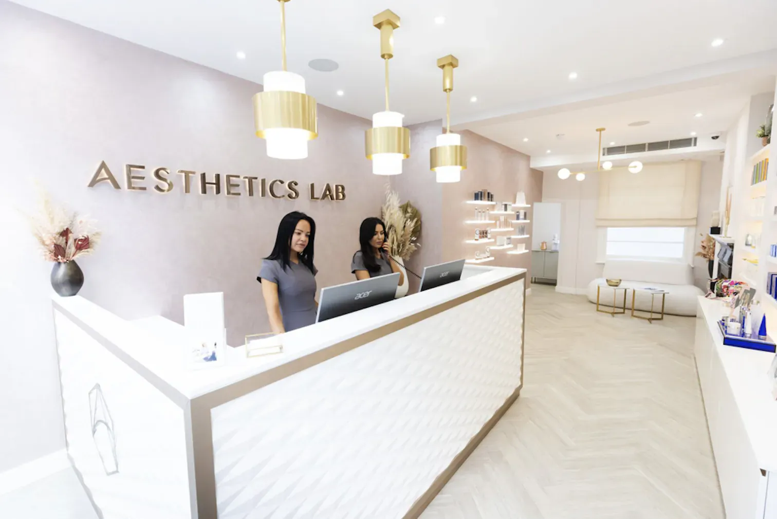 Two women at the white reception desk of Aesthetics Lab, modern clinic with gold pendant lights and skincare products.