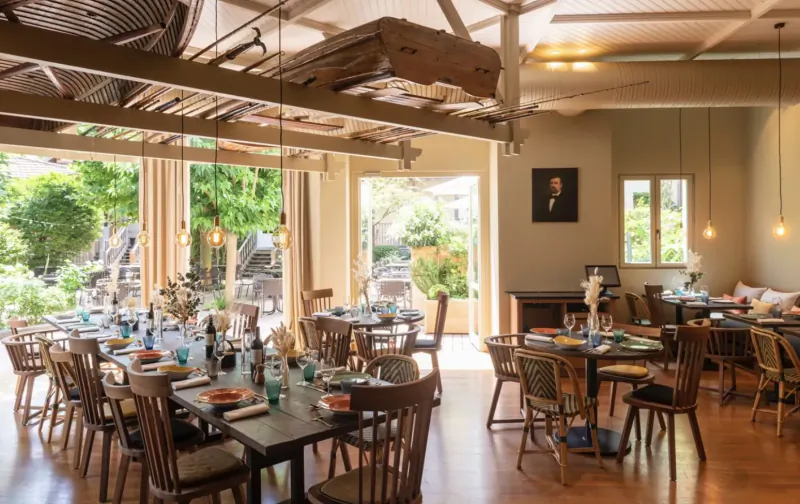 Elegant dining room at Les Étangs de Corot with wooden tables set for meals, large windows to lush garden, warm lighting.