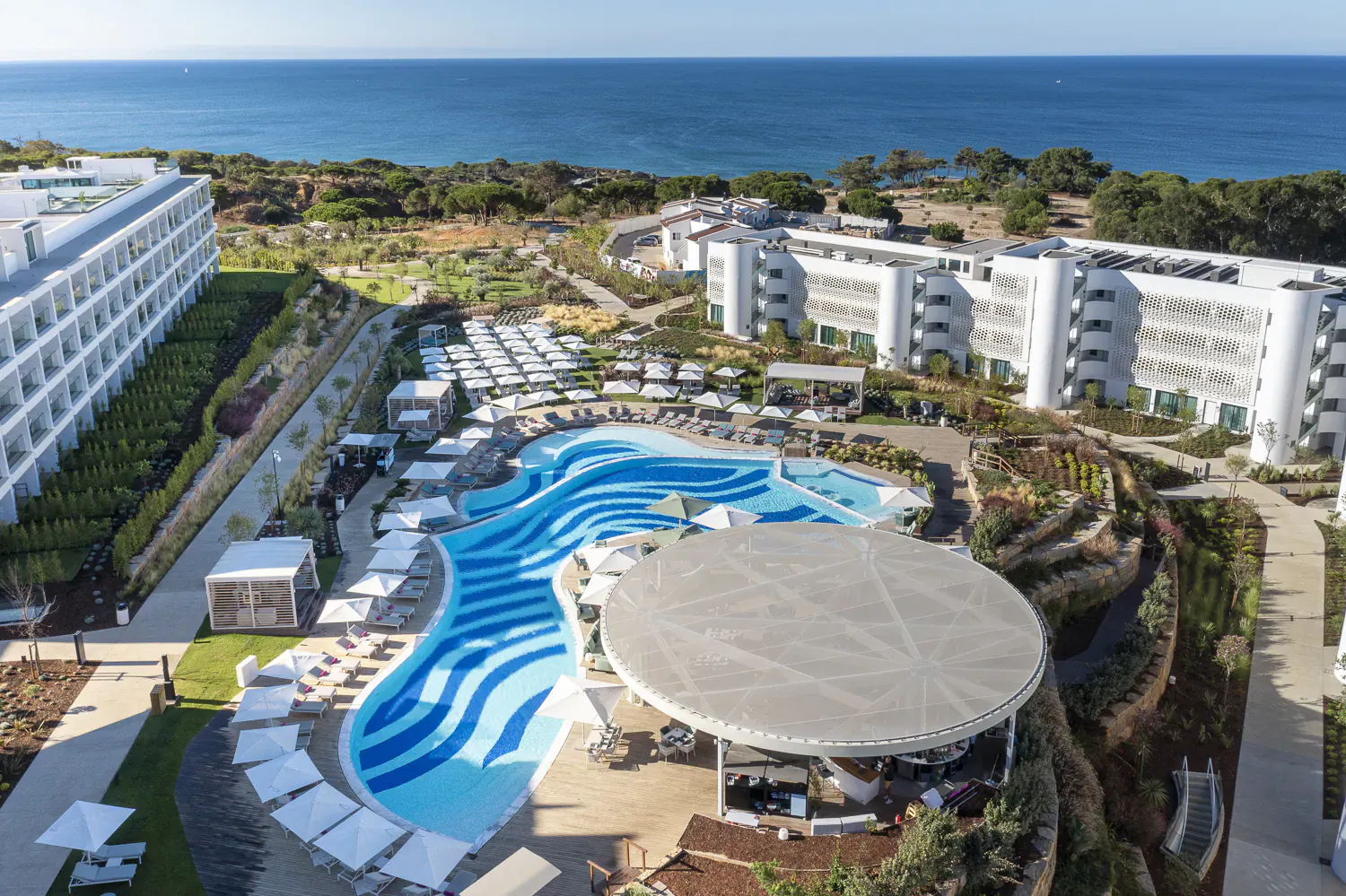 Aerial view of W Algarve Hotel's wavy blue infinity pool, loungers, cabanas, white buildings, and ocean on Portugal's coast.