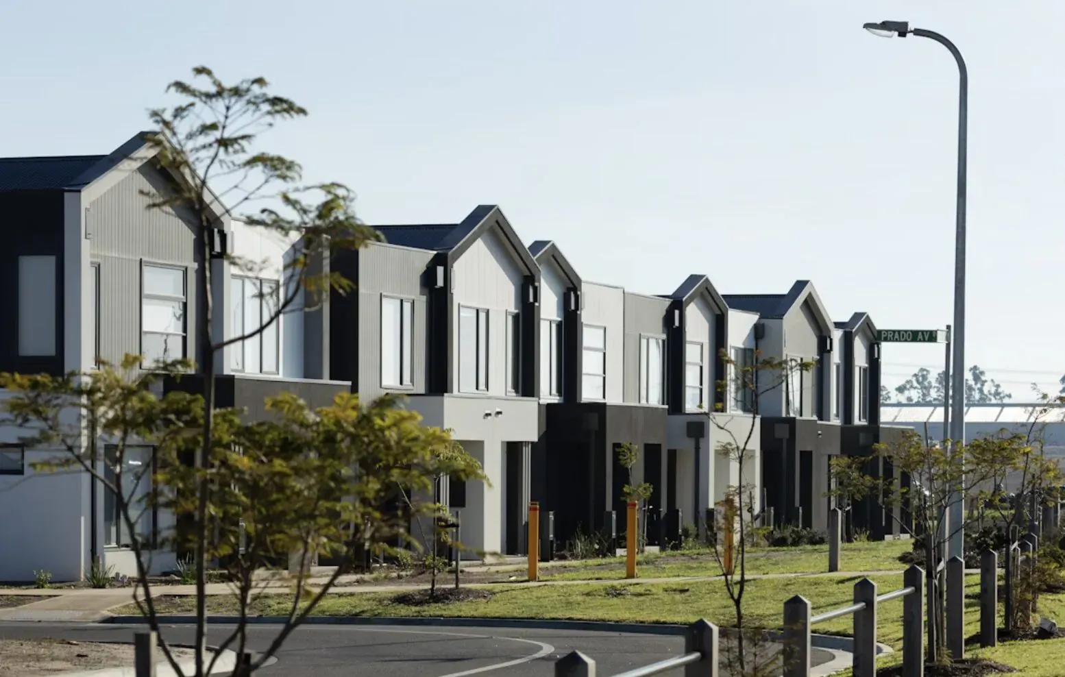 Row of modern grey and white townhouses with trees, streetlights, and fence on sunny day