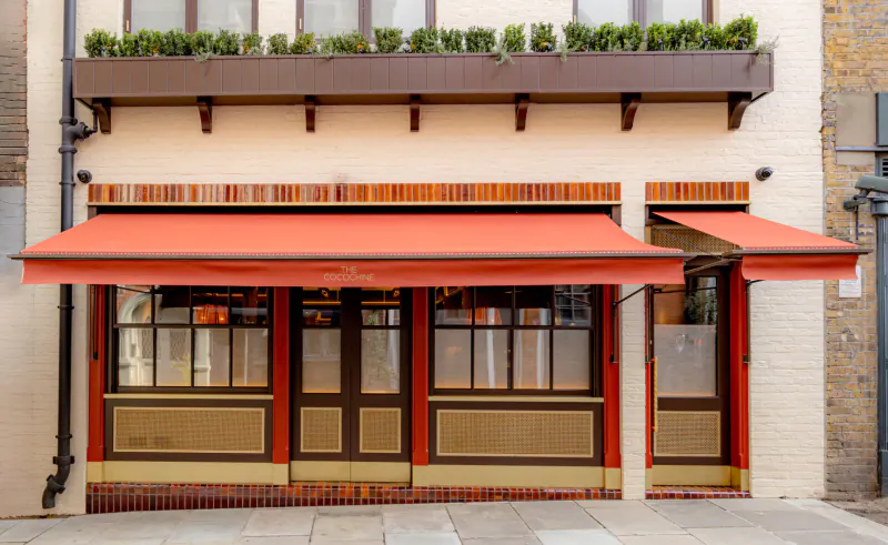 The Cocochine restaurant facade with orange awning, large windows, plants in window boxes, and beige brick exterior on city street.