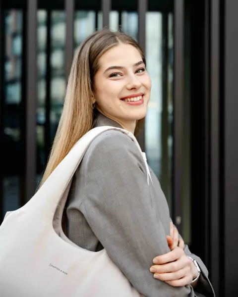 Smiling young woman with long blonde hair in gray blazer and white Kapten & Son tote bag, standing by metal gate.