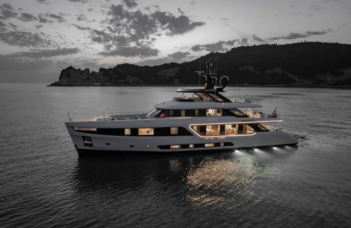 Nomen Omen, sleek gray Benetti superyacht on calm sea at twilight with rocky cliffs backdrop
