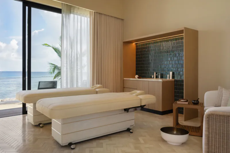 Luxurious spa room with two white massage tables, wooden cabinet, and ocean view through large windows at Maldives resort.