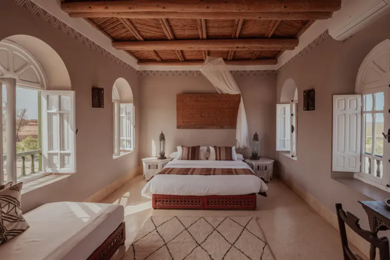Spacious Moroccan bedroom in Essaouira villa with wooden ceiling, king bed, mosquito net, arched windows, and rugs.