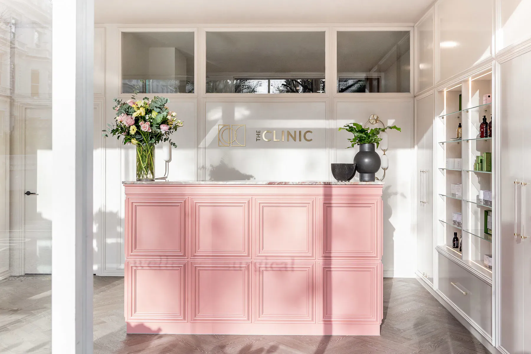 Pink panelled reception desk at The Clinic with yellow flowers, plants, and products on white shelves.