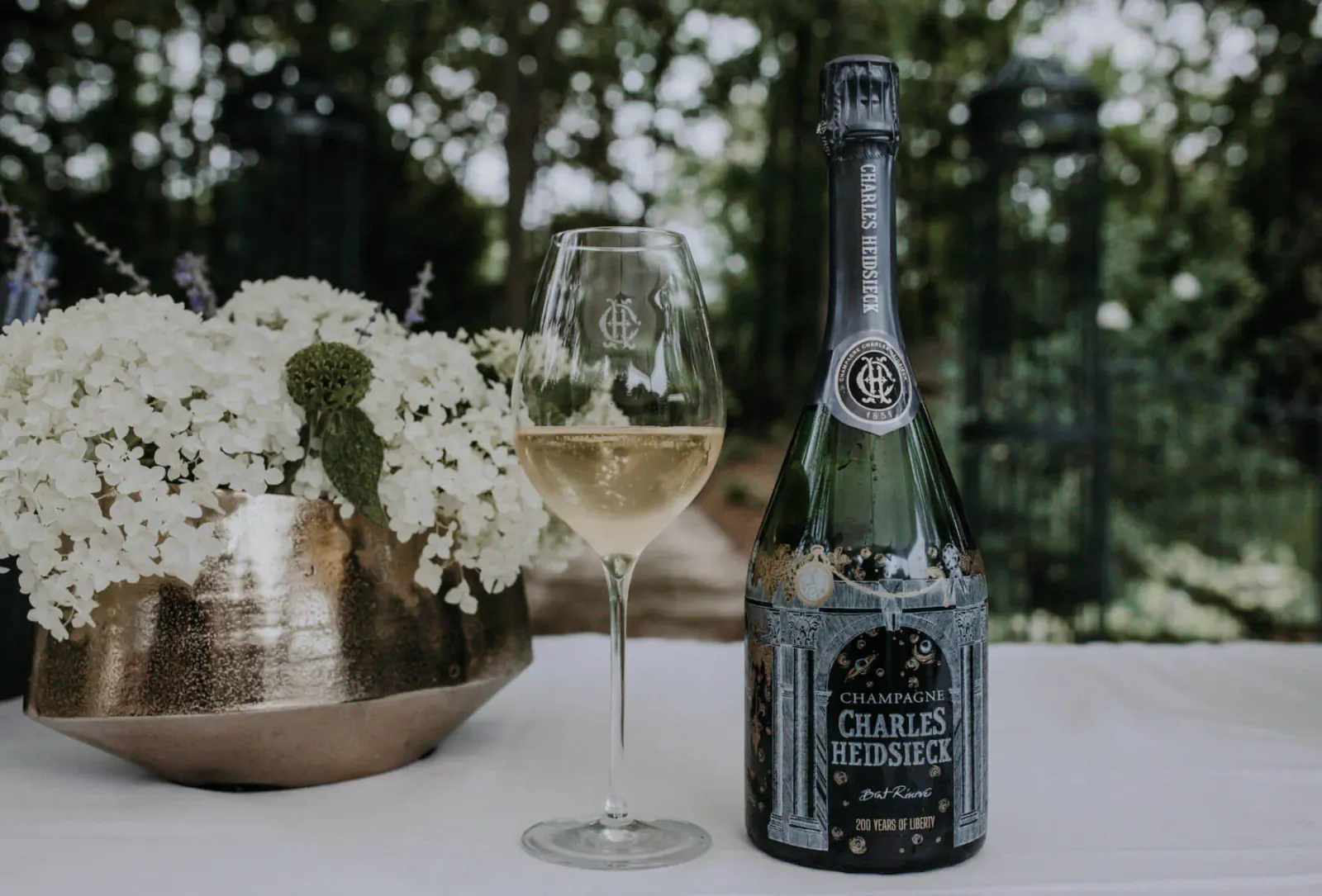 Charles Heidsieck 200th Anniversary champagne bottle with poured glass, white flowers, garden backdrop