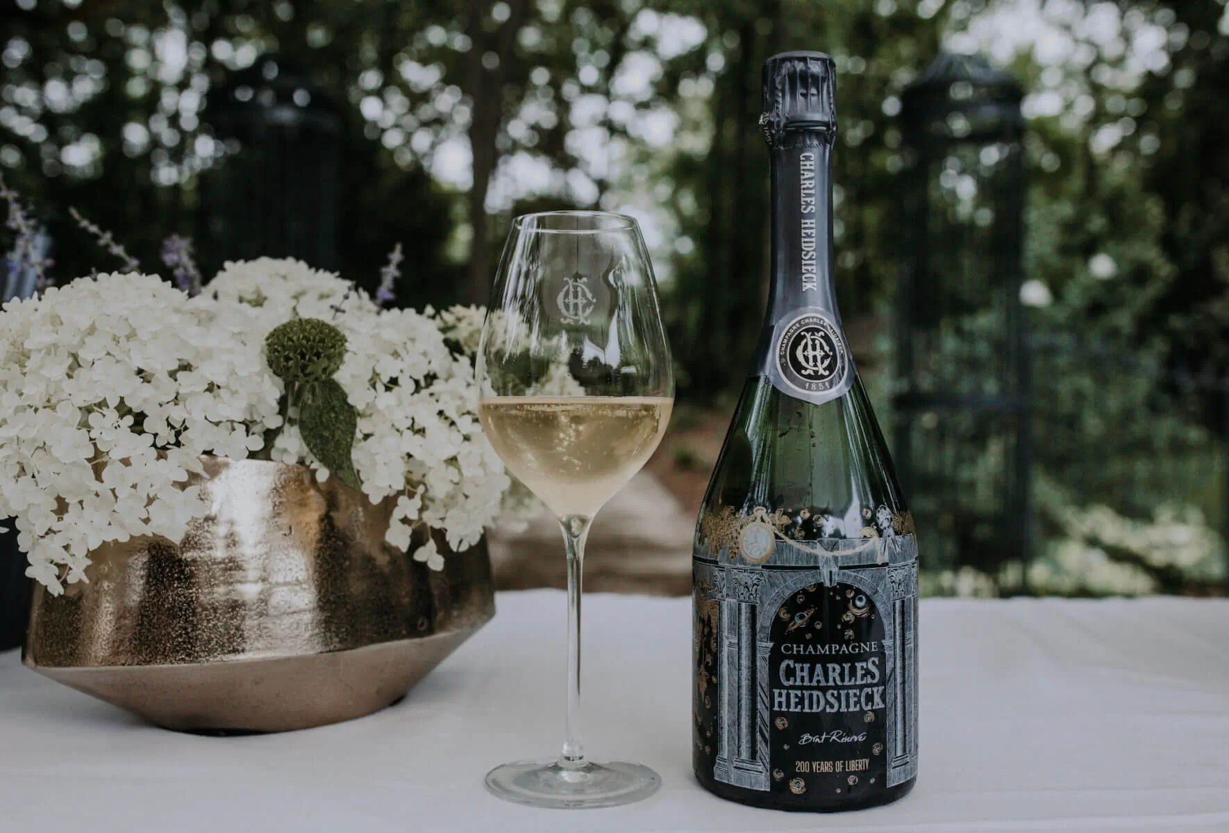 Charles Heidsieck 200th Anniversary champagne bottle with poured glass, white flowers, garden backdrop