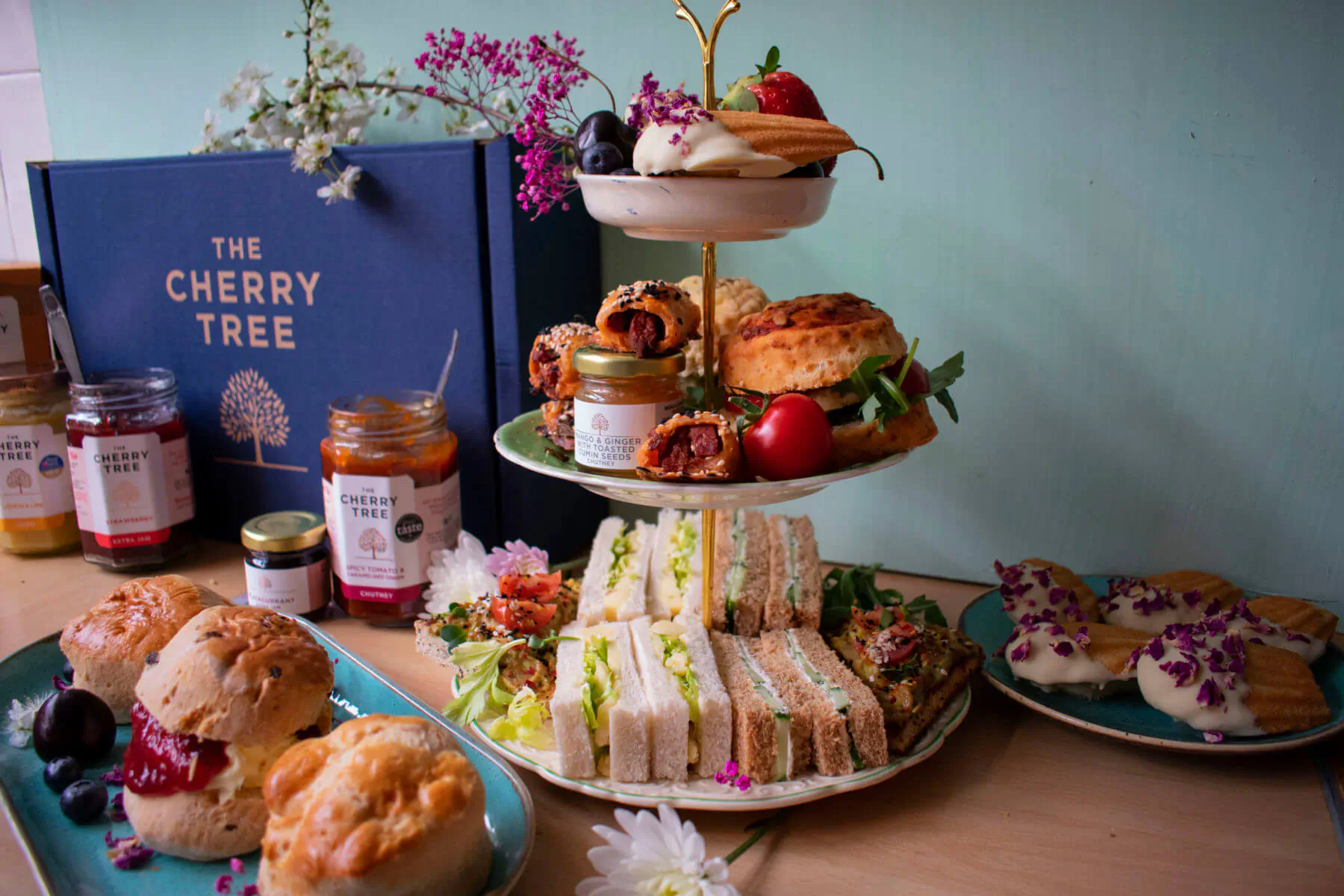 The Cherry Tree artisan afternoon tea on a three-tier stand with scones, sandwiches, cakes, jam jars, and flowers beside a branded box