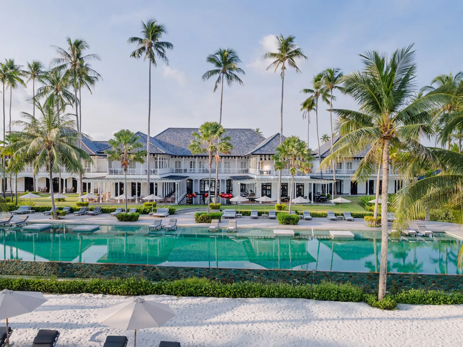 The Sanchaya luxury resort with white villas, tall palms, large green pool, and white sand beachfront.