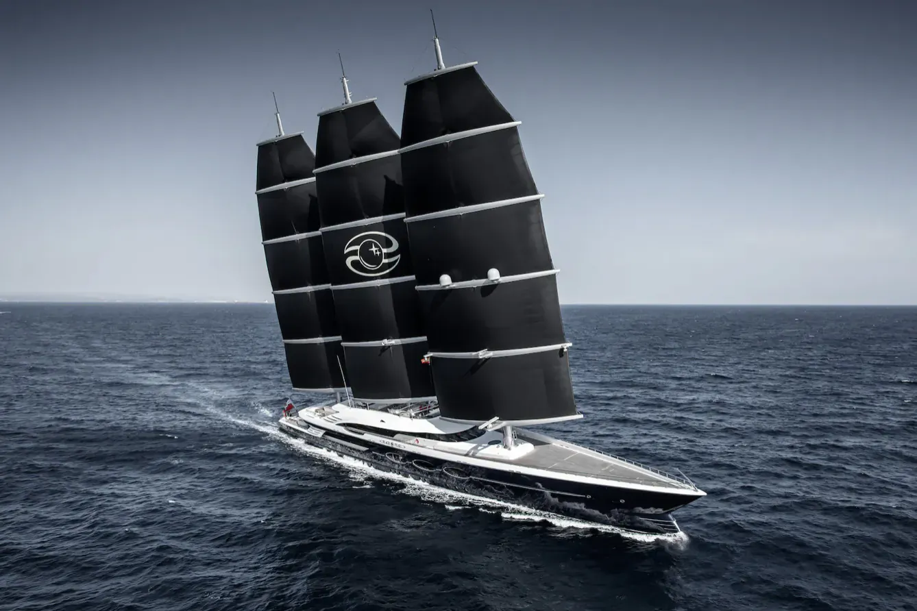Sleek black luxury superyacht with three tall black sails and circular logo, sailing on ocean.