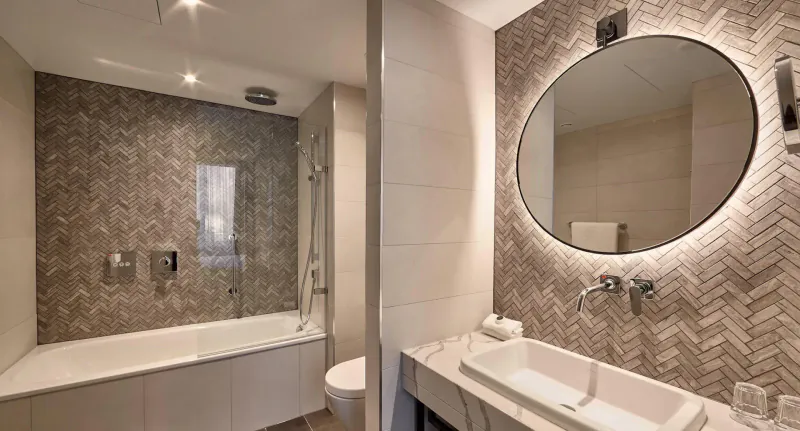 Modern luxury bathroom in Fellows House Cambridge apartment with glass shower, bathtub, oval mirror, and herringbone tiles.