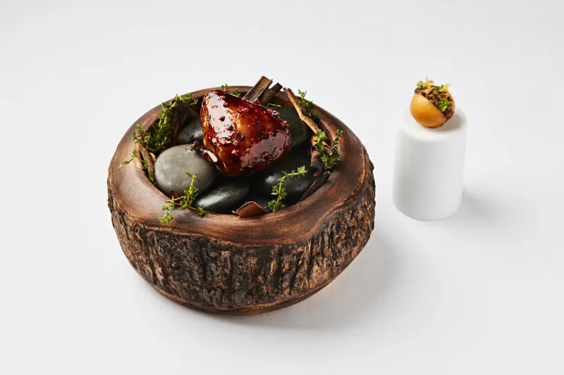 Glazed pear in wooden bowl with moss, stones, cinnamon stick; beside white cylinder and orange chestnut sphere