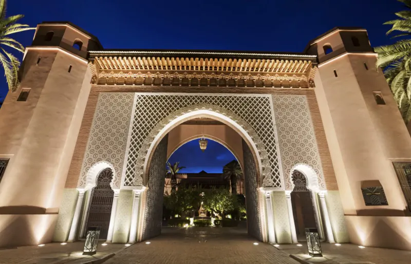 Lit Moorish archway entrance to Royal Mansour Marrakech medina at night, flanked by palms and gardens.