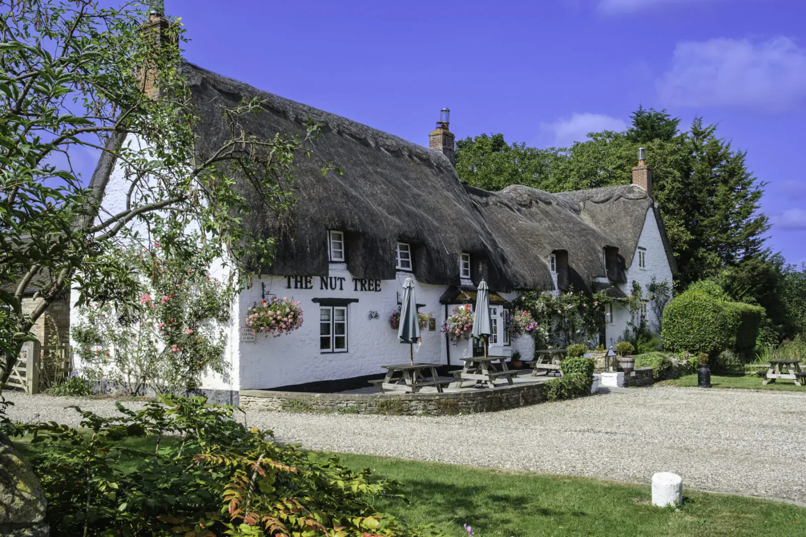 The Nut Tree Inn, thatched-roof pub with flower baskets and umbrellas, on gravel courtyard under blue sky.