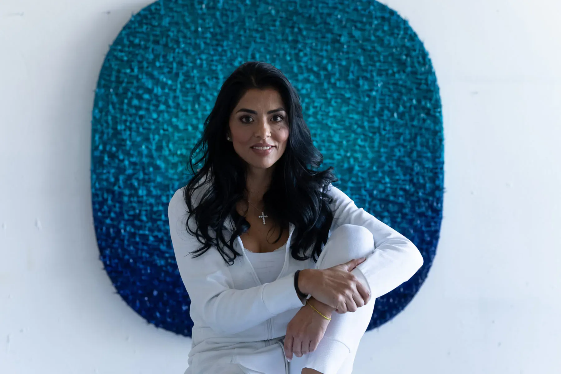 Artist Susana Anaya smiles, seated in white top with legs crossed before teal mosaic wall