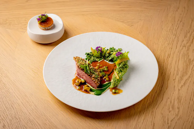Elegant plated dish of seared duck breast with orange sauce, greens, edible flowers, and side beet tartlet on white plate.