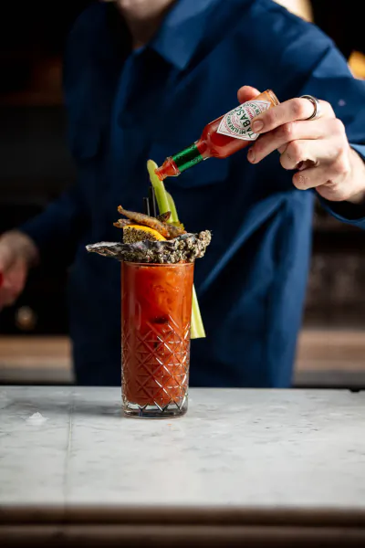 Bartender in blue shirt pours Tabasco sauce from green-topped bottle onto garnished Bloody Mary cocktail on bar counter.