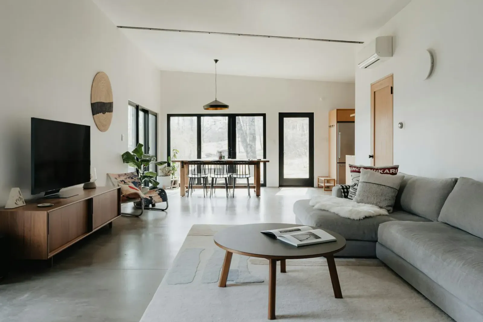 Modern open-plan living room with gray L-shaped sofa, wooden coffee table, TV stand, dining table, and plants.