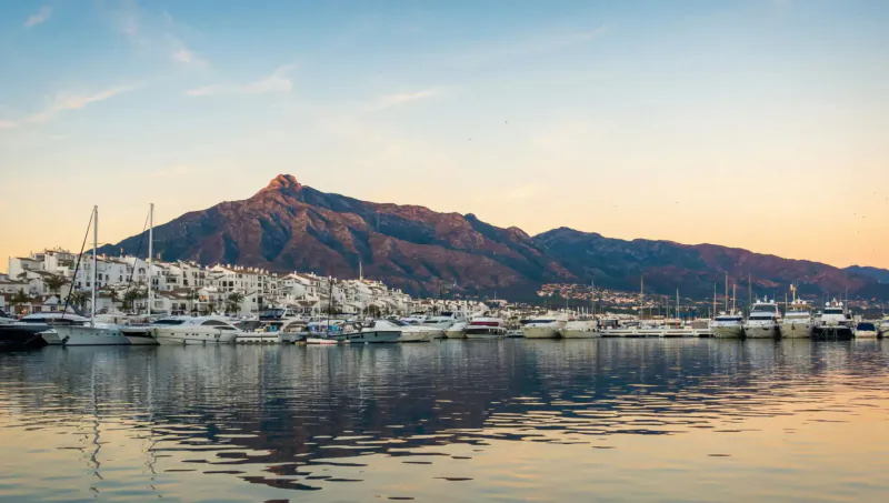 Sunset view of Marbella marina with white yachts, luxury villas, and rugged mountains reflecting in calm water