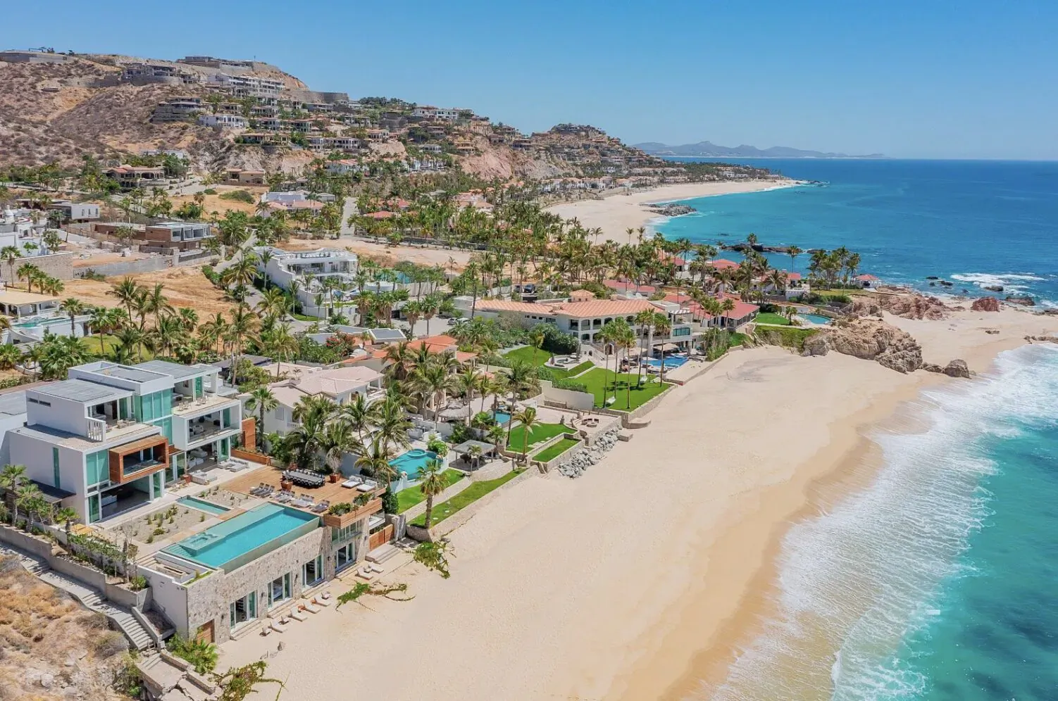 Aerial view of luxury villas and homes on a sandy beach with turquoise ocean waves in Cabo San Lucas.