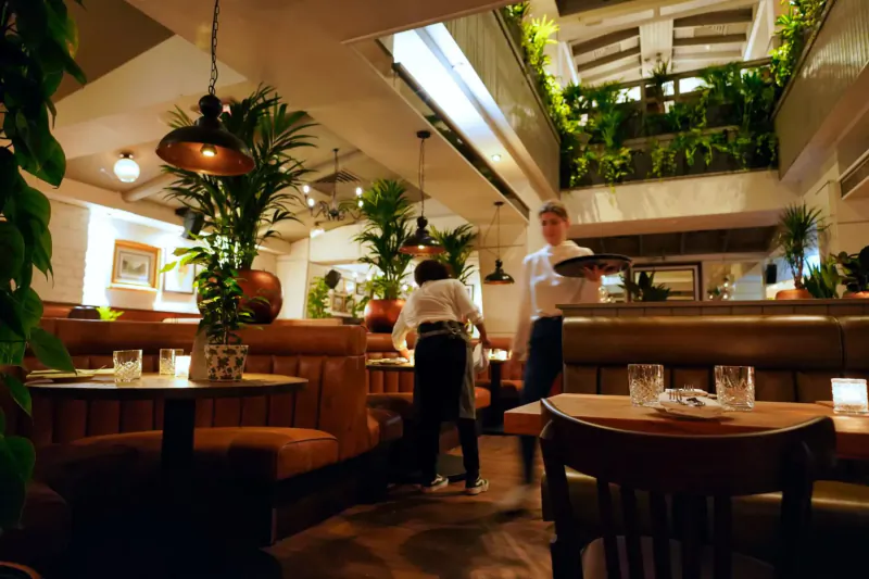 Busy upscale restaurant interior with servers at leather booths, plants, warm lighting, and greenery atrium