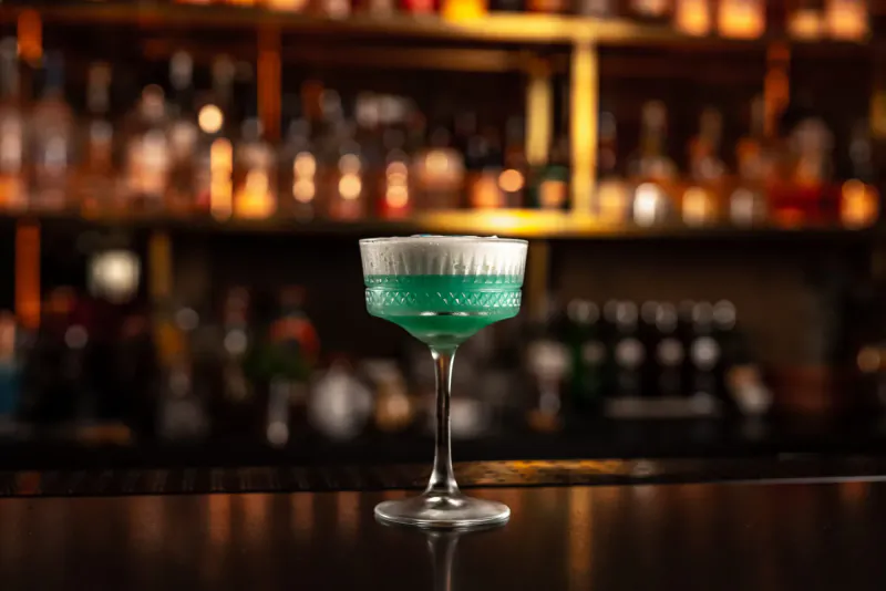 Green cocktail in coupe glass with white foam top, on bar counter with blurred golden shelves of bottles behind.