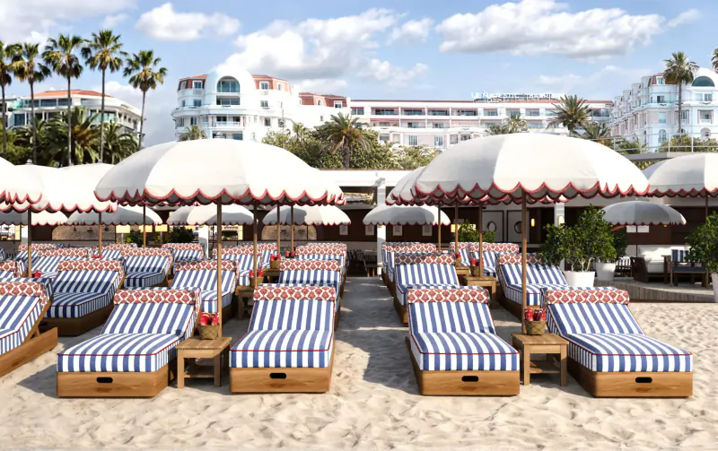 Luxury beachside lounge at Hotel Barrière Le Majestic Cannes with white umbrellas, blue-striped teak loungers, palm trees, and white hotel buildings.