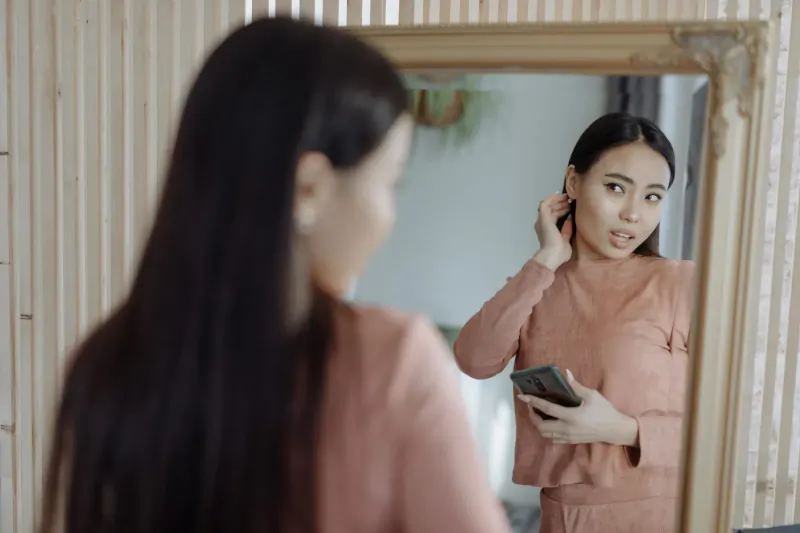 Asian woman in elegant peach loungewear checks earrings in ornate mirror, holding phone, relaxed indoor setting