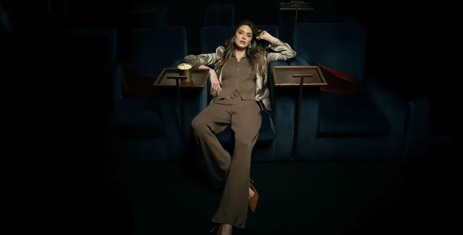 Woman in brown lounge suit reclining relaxed in dark cinema theater seats with tray tables
